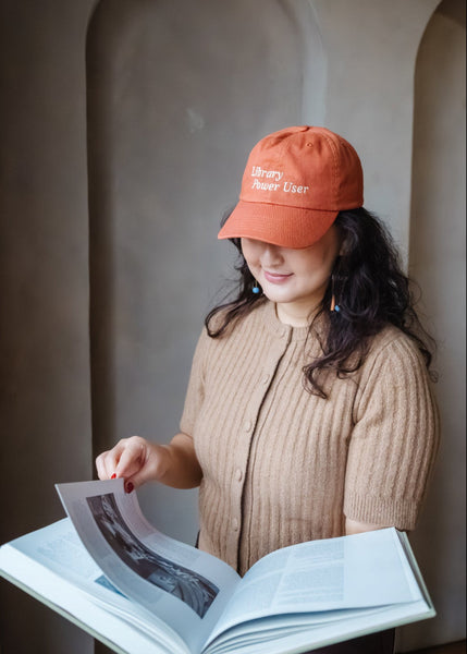 Woman wearing a terra cotta colored dad hat with 'Library Power User' text, holding an open book against a neutral wall.