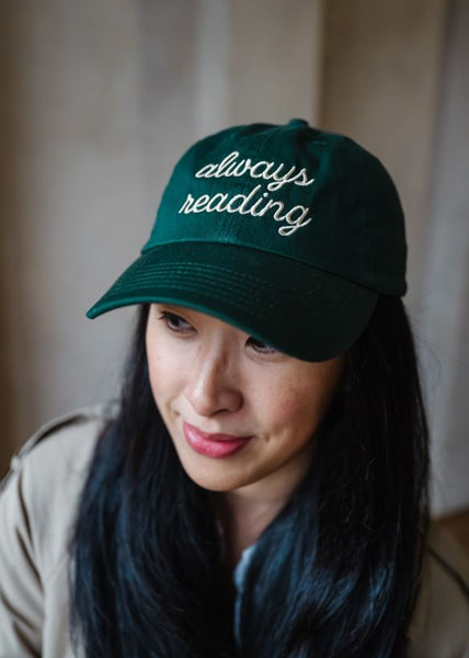 Woman wearing a dark green cap with 'always reading' embroidered text against a neutral background